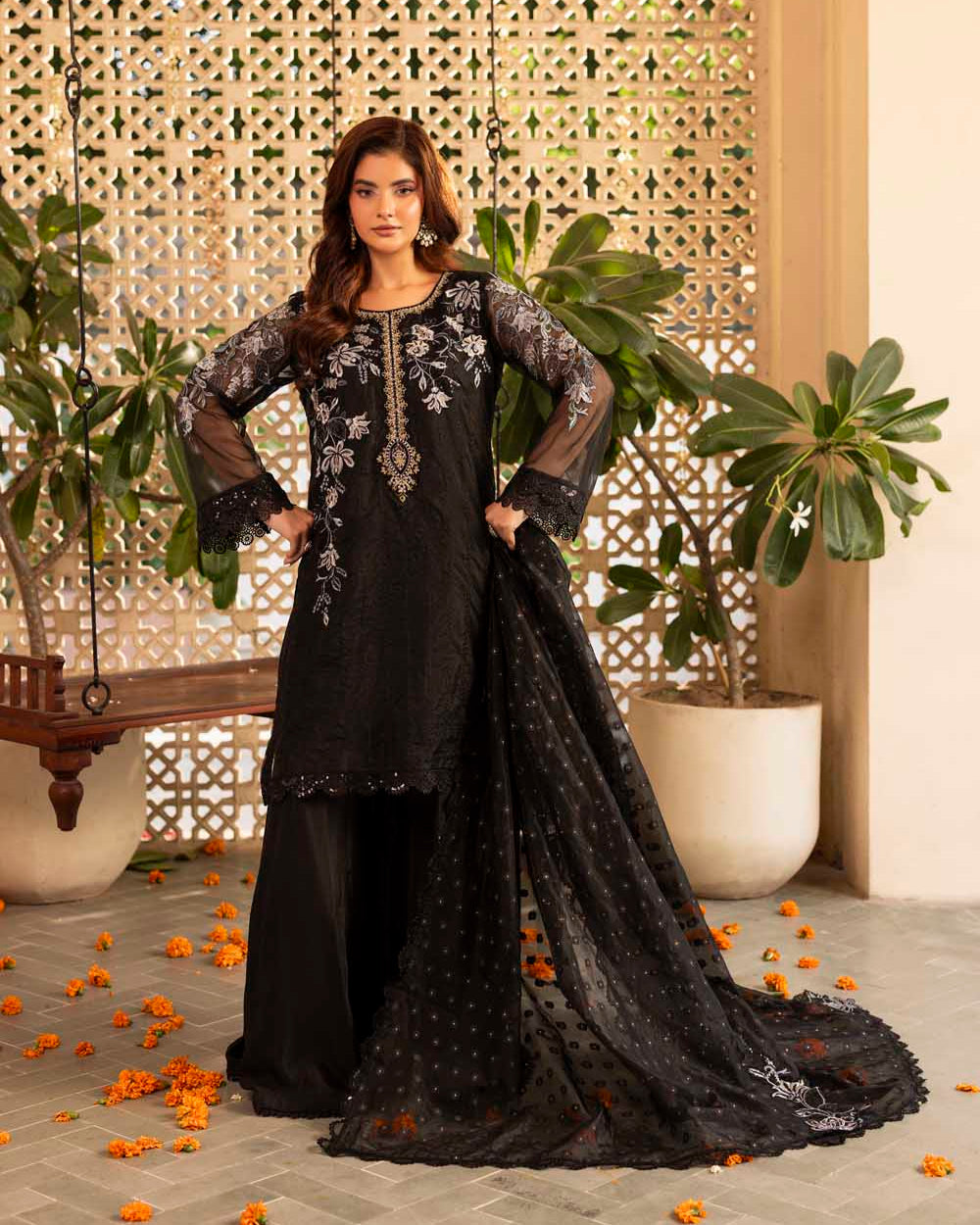 Woman in a black embroidered outfit standing in a decorative indoor setting with plants and a lattice wall.