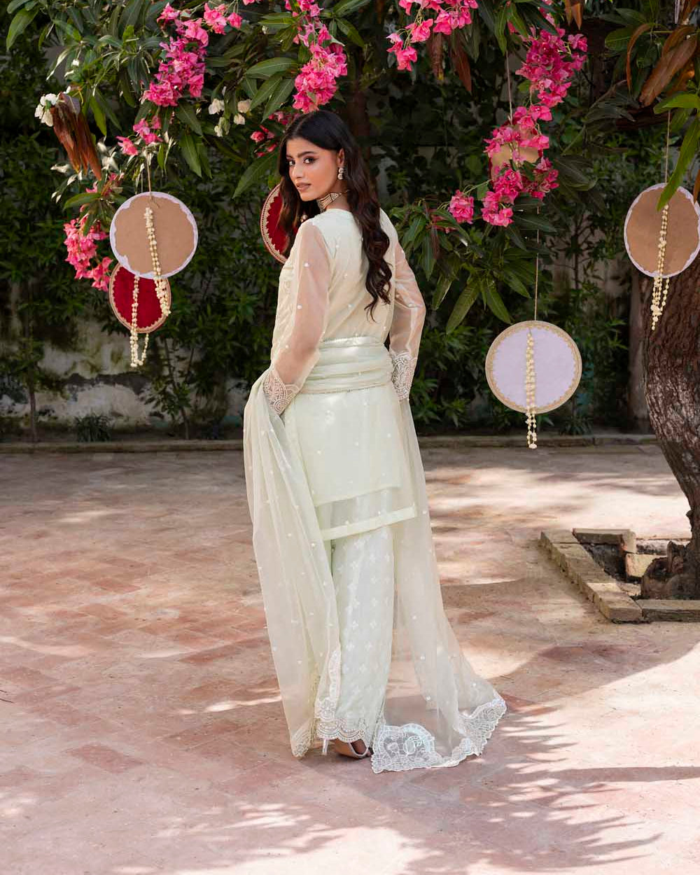 Woman in a white traditional outfit standing under a floral arch with pink flowers.