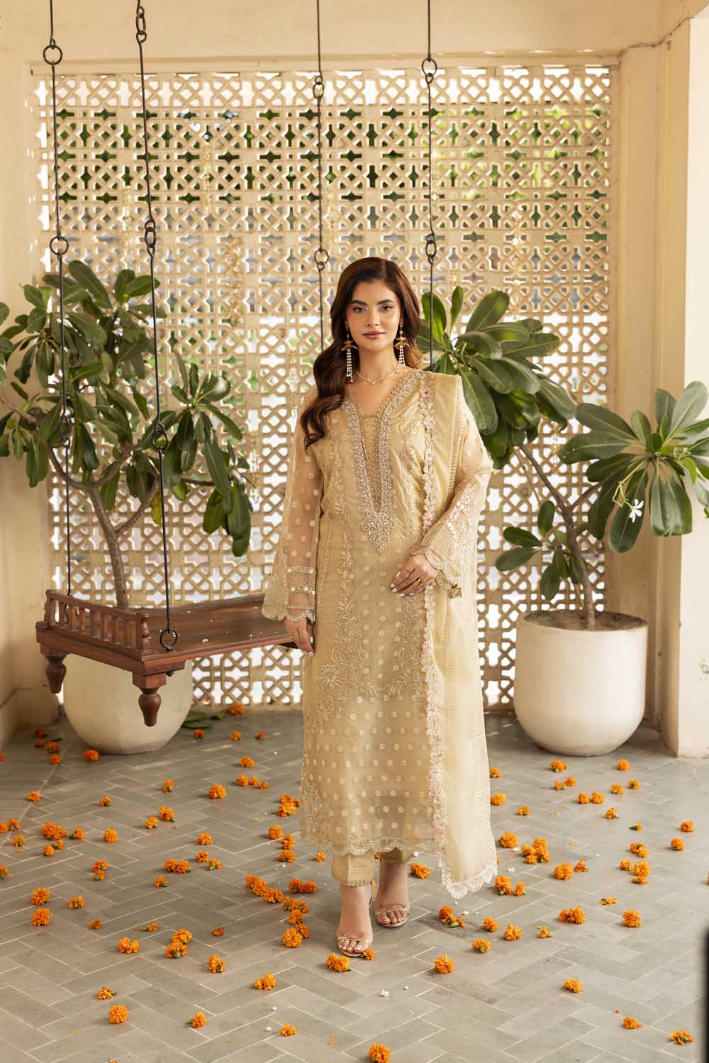 Woman in a beige traditional outfit standing in a decorated indoor setting with plants and decorative elements.