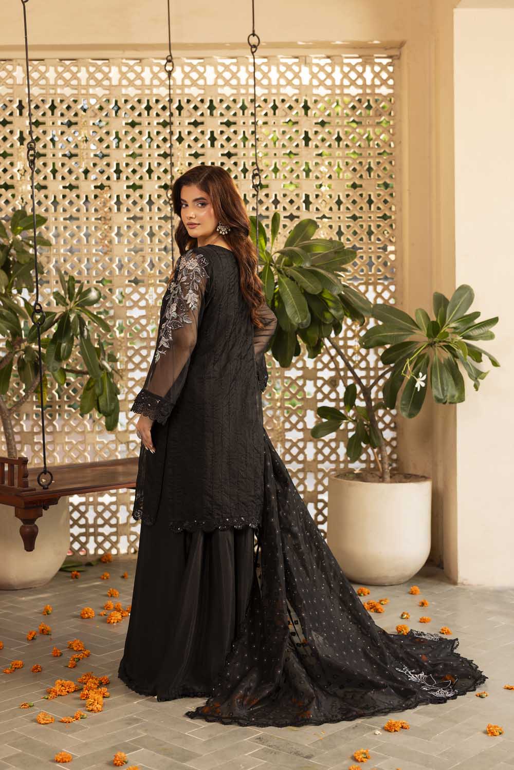Woman in a black traditional outfit standing in a decorative indoor setting with plants and a lattice wall.
