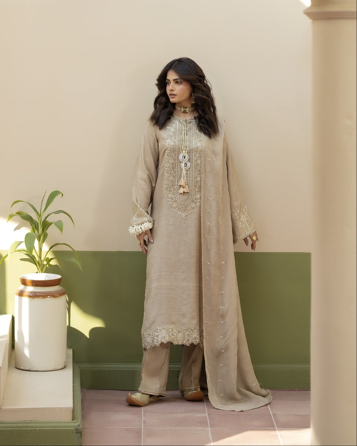 Woman in a beige embroidered outfit standing in a sunlit room with green walls and a plant.