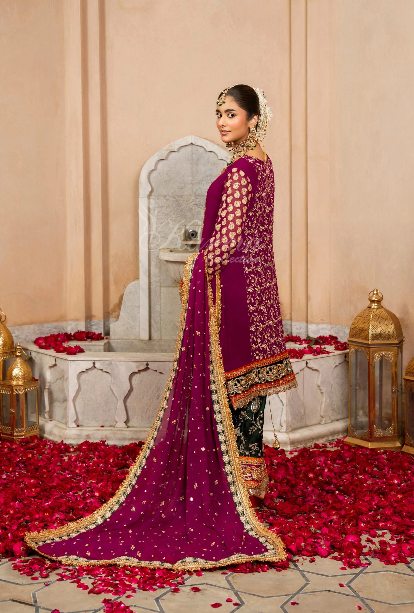 Woman in a traditional purple saree with gold details standing in a decorated room with red flowers and lanterns.