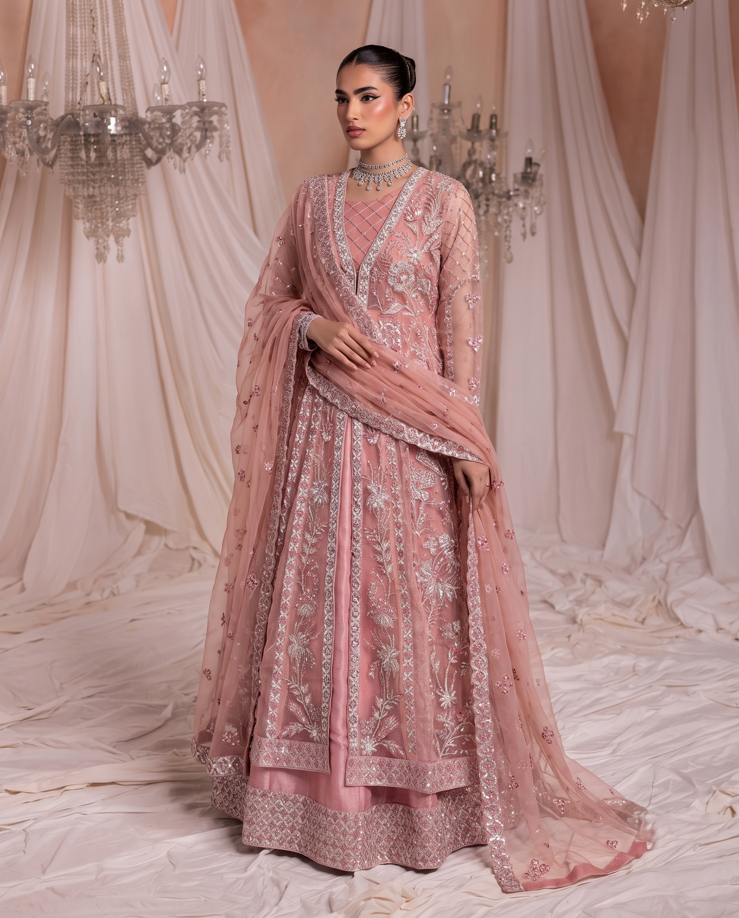 A woman models a dusty pink embellished traditional outfit with a matching dupatta, set against a draped cream fabric backdrop with hanging crystal chandeliers.