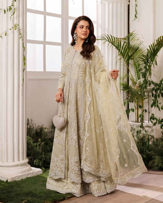 A woman models a traditional South Asian ensemble featuring a heavily embroidered light beige/cream kurta and matching dupatta, styled with a small heart-shaped clutch in an elegant indoor garden setting with white pillars.
