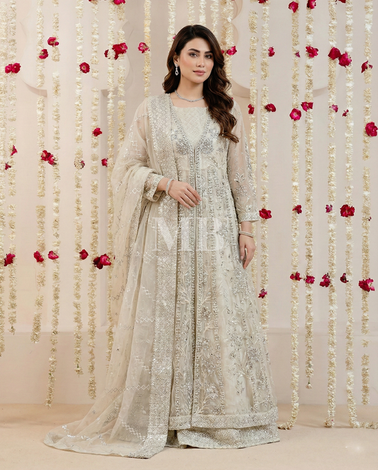 Woman in a light-colored embroidered traditional outfit standing against a decorative floral and string backdrop.
