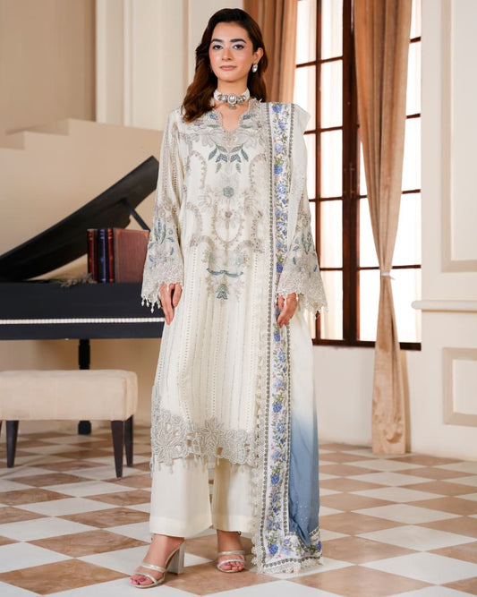 A woman models a traditional South Asian outfit in a grand interior room with a checkered floor, a grand piano, and tall curtains. The outfit is cream-colored with intricate silver embroidery and a blue gradient dupatta.