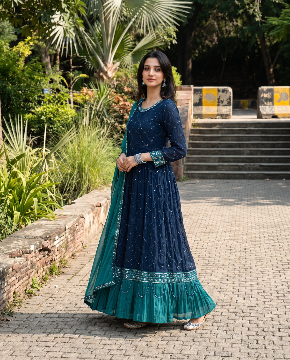 A woman stands outdoors on a paved walkway wearing a dark teal blue traditional long dress with a matching teal dupatta. The dress features silver mirror work embroidery on the bodice and a tiered ruffled hem with a decorative border.