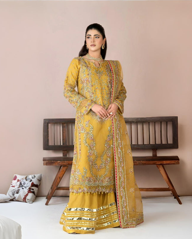Woman wearing a yellow embroidered traditional outfit in a room with a wooden bench and decorative pillows.