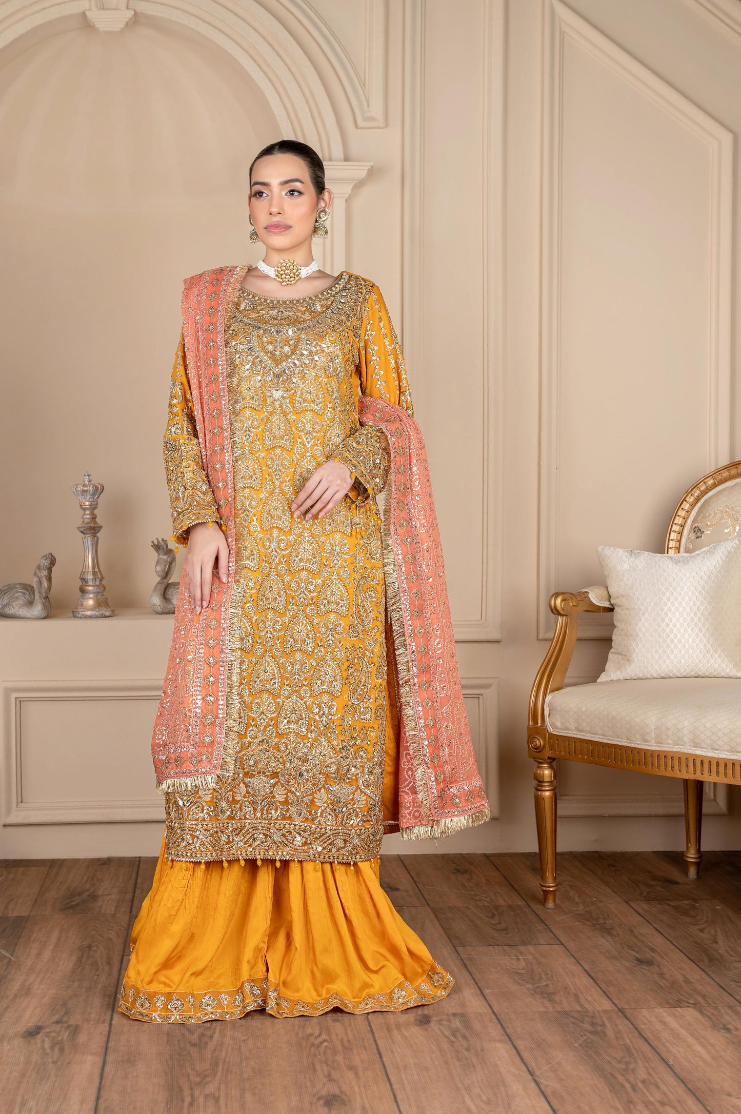 Woman in a yellow and gold embroidered traditional outfit with a pink dupatta in an elegant room.